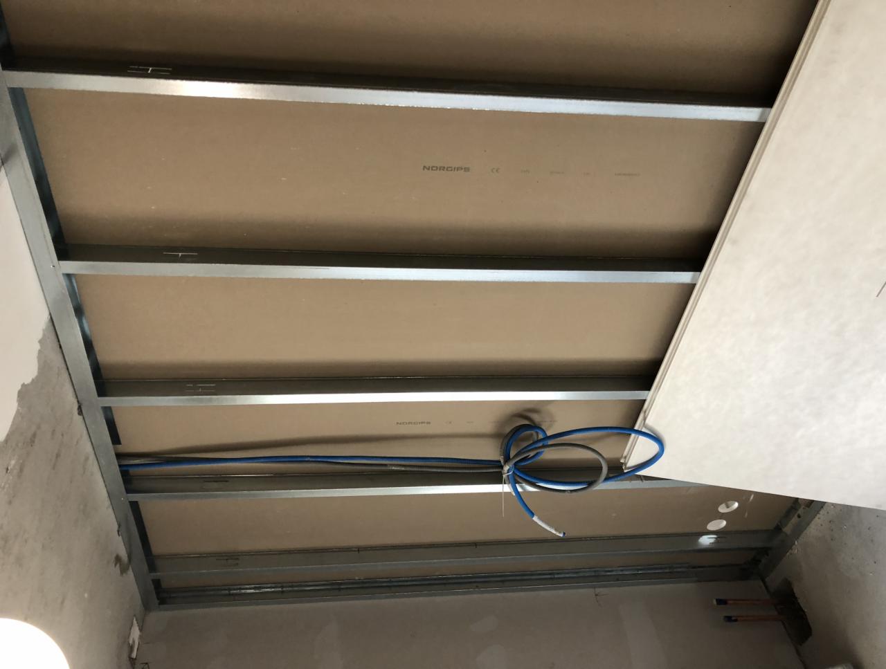 Konstrukcja sufitu podwieszanego z profili metalowych i częściowo zamontowanych płyt. Widoczne przewody elektryczne. Nierówna powierzchnia ściany w tle.