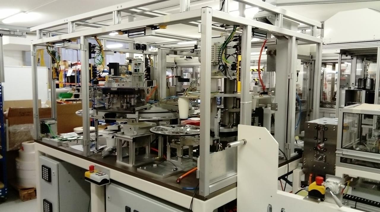 Kompleksowa linia produkcyjna z aluminiową ramą, widoczne elementy automatyki, przewody i panele sterowania, w tle magazynowe kartony.