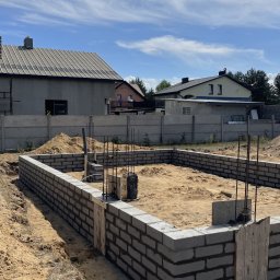 Polbudrol - Generalny Wykonawca - Fundamenty domu w trakcie budowy: szare bloczki betonowe, wystające pręty zbrojeniowe, piasek. W tle domy jednorodzinne i betonowe ogrodzenie.