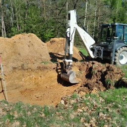 Polbudrol - Generalny Wykonawca - Wykop pod fundamenty w Starachowicach: pracownik z łatą geodezyjną i koparka Terex na tle drzew. Realizacja budowy domu w stanie surowym otwartym.