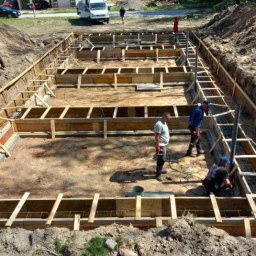 Polbudrol - Generalny Wykonawca - Budowa fundamentów domu jednorodzinnego: szalunek z drewna, zbrojenie stalowe. Na dalszym planie budynek w stanie surowym otoczony zielenią.