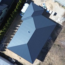 Polbudrol - Generalny Wykonawca - Zdjęcie z drona przedstawia ukończony dach z blachy na domu w stanie deweloperskim. Widoczne elementy konstrukcyjne, okolica i materiały budowlane.
