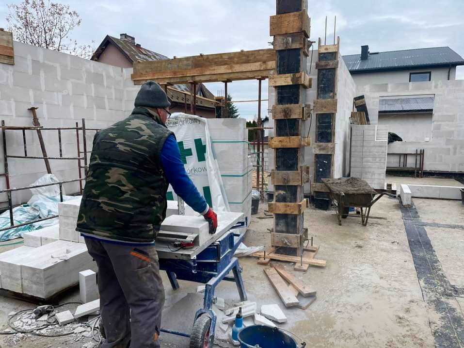 Pracownik w kamizelce moro tnie bloczek z betonu komórkowego na niebieskiej pile stołowej na budowie domu murowanego z betonu komórkowego; w tle widoczne szalunki kolumn, taczka z zaprawą i domy...