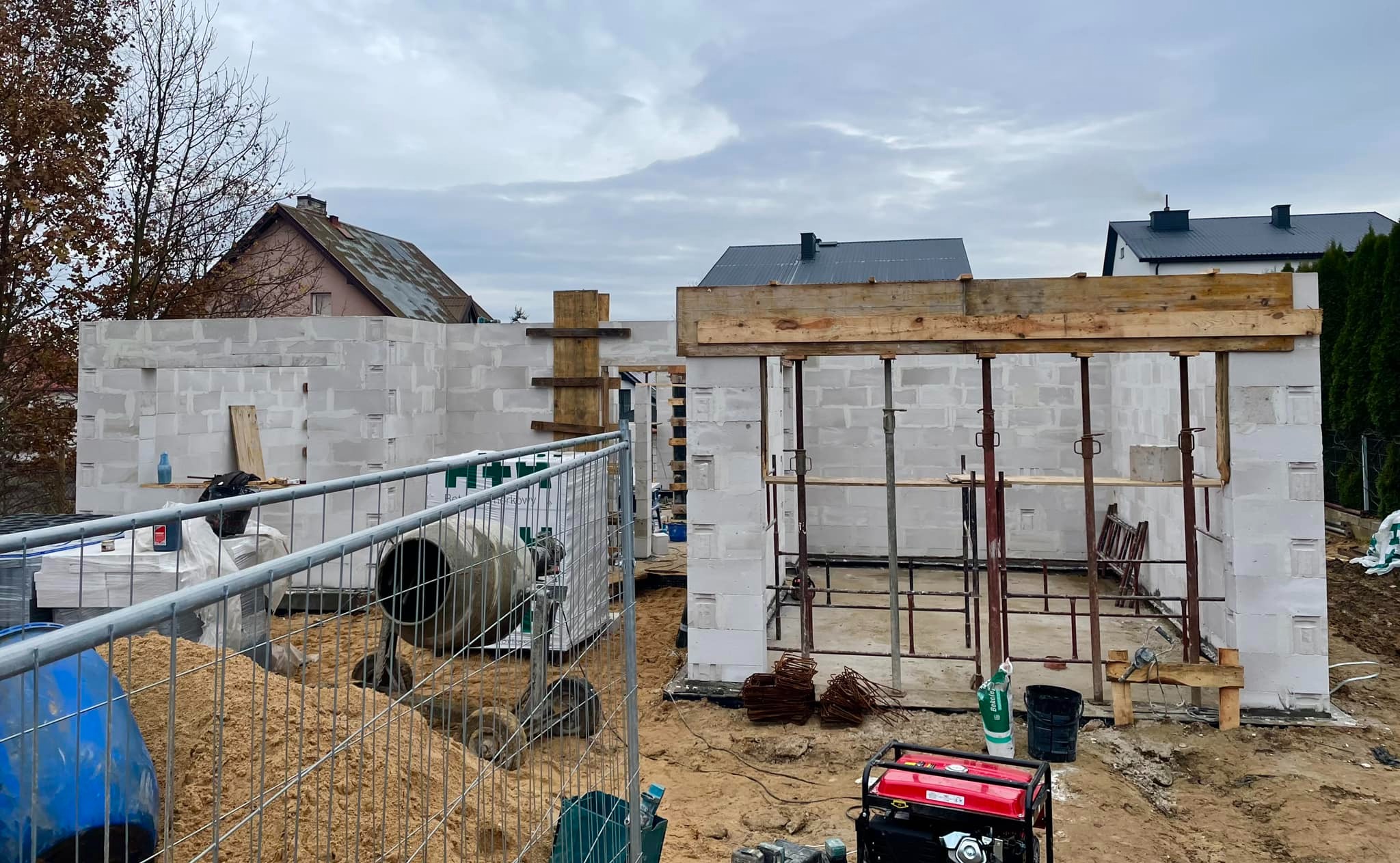 Budowa domu z białych bloczków: widoczne ściany w stanie surowym, betonowa mieszarka, generator prądu, stal zbrojeniowa, drewniane szalunki oraz materiały budowlane za ogrodzeniem placu budowy.