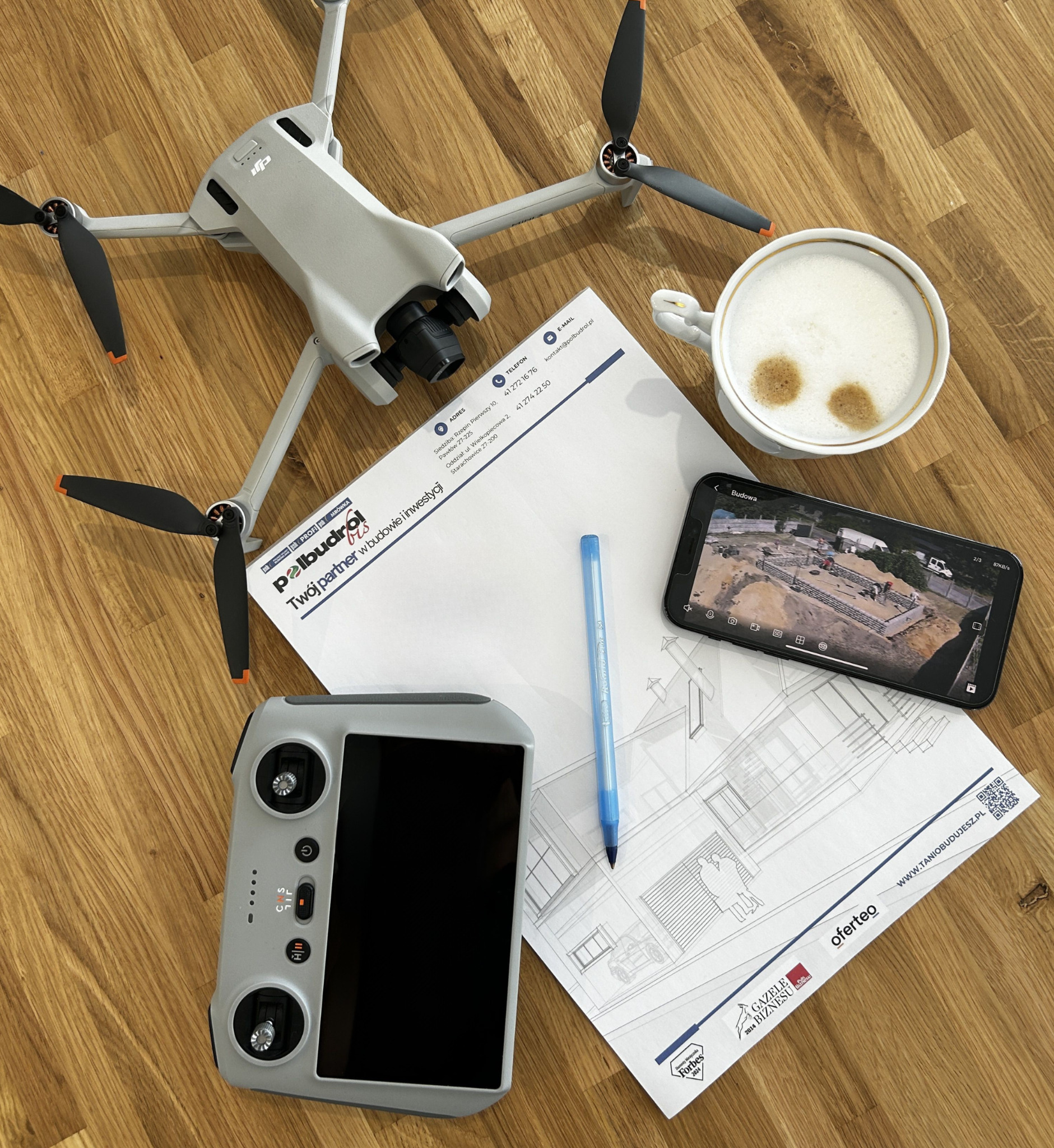 Dron DJI Mini 3 Pro, kontroler, telefon z podglądem budowy, projekt domu i kawa na biurku. Nowoczesne narzędzia w budownictwie.