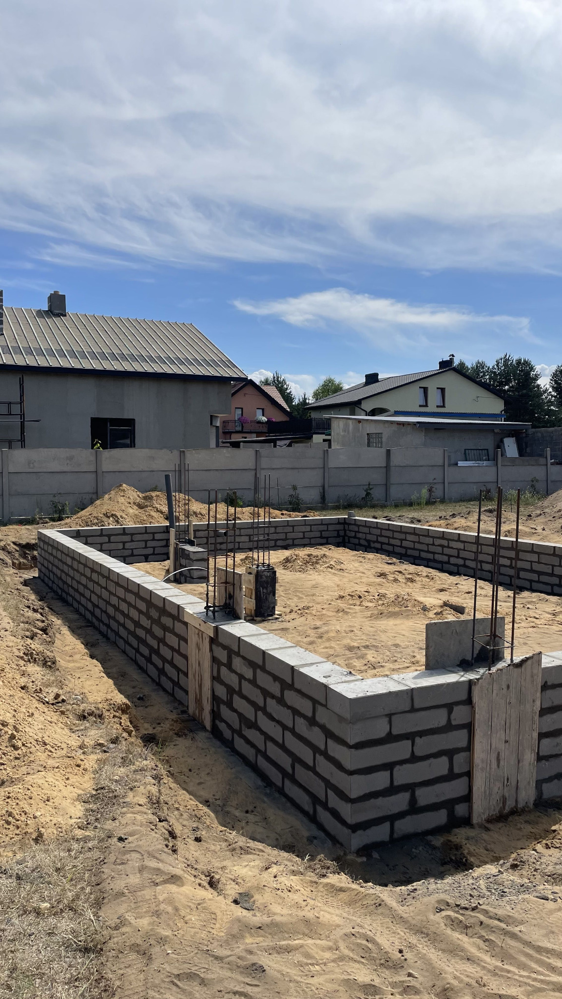 Fundamenty domu w trakcie budowy: szare bloczki betonowe, wystające pręty zbrojeniowe, piasek. W tle domy jednorodzinne i betonowe ogrodzenie.