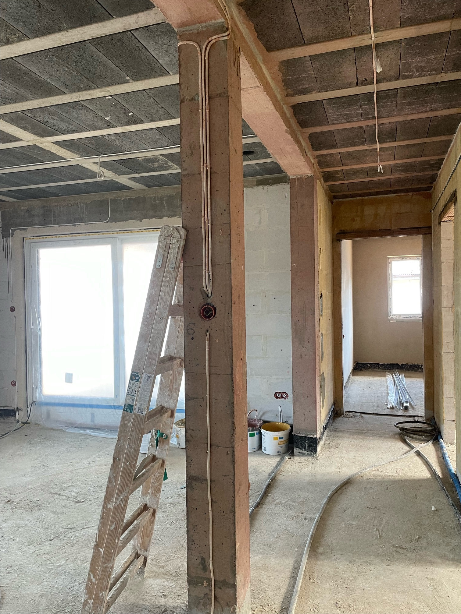 Wnętrze w trakcie budowy: widok na surowy strop, instalacje elektryczne w ścianach, okno zabezpieczone folią i drewnianą drabinę opartą o filar. Stan deweloperski.