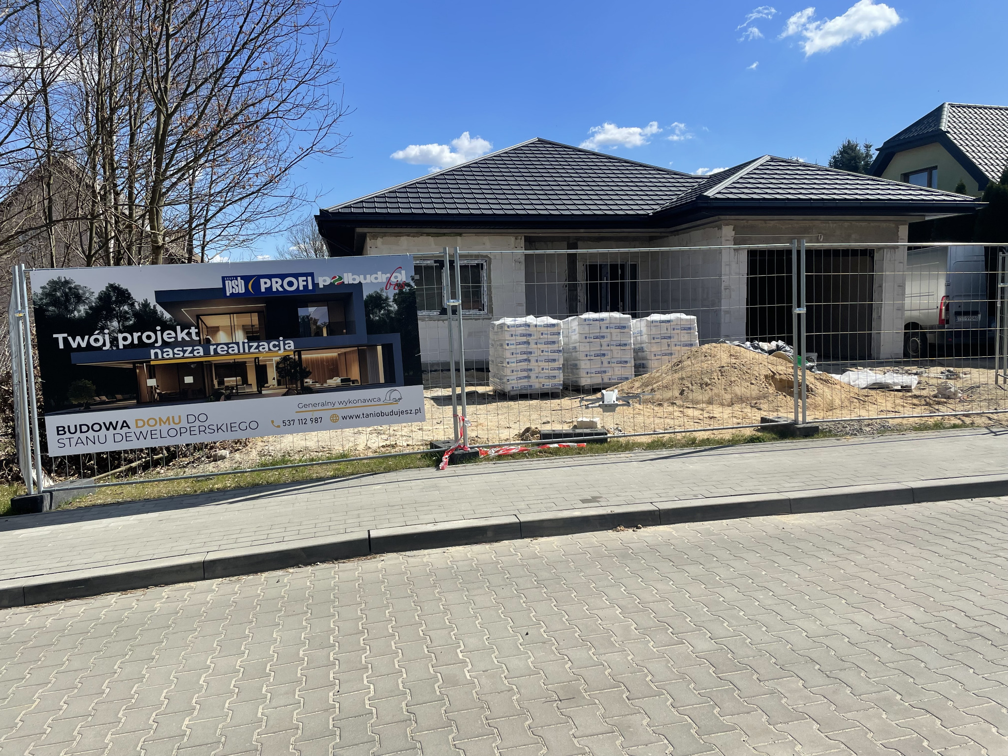 Budowa domu parterowego do stanu deweloperskiego, widoczna zza ogrodzenia. Na pierwszym planie reklama firmy budowlanej z wizualizacją projektu.