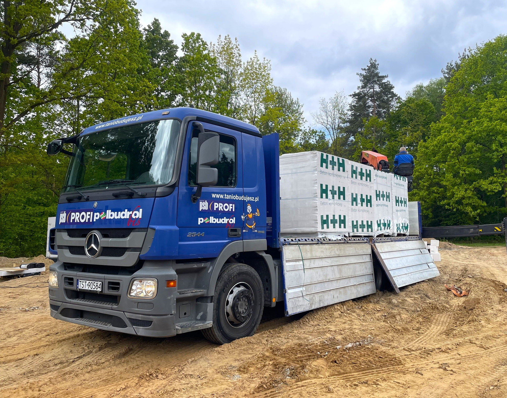 Niebieska ciężarówka z dźwigiem HDS, załadowana bloczkami H+H, stoi na placu budowy. Widoczny pracownik na górze ładunku i logo firmy 'PROFI pelbudrol'.