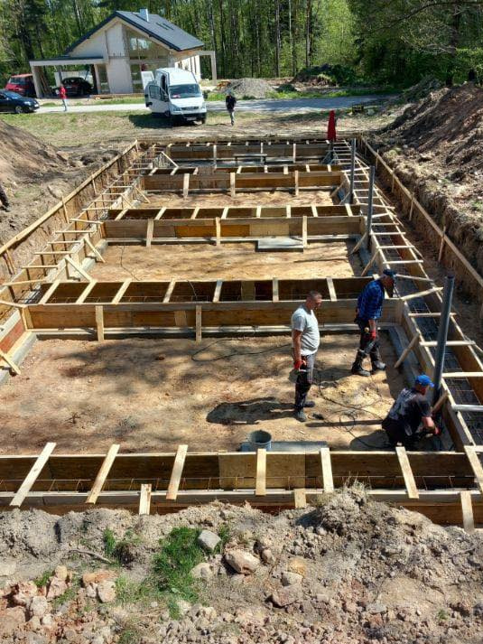Budowa fundamentów domu jednorodzinnego: szalunek z drewna, zbrojenie stalowe. Na dalszym planie budynek w stanie surowym otoczony zielenią.