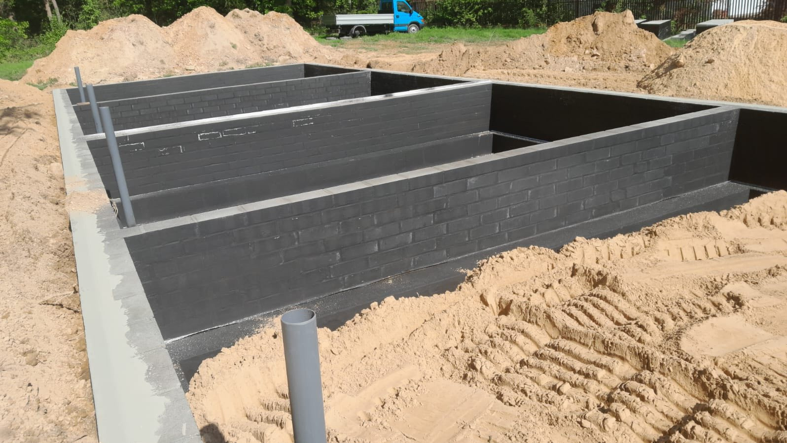 Fundamenty domu w trakcie budowy: czarne bloczki fundamentowe, szalunek z betonu, rury pionowe, piasek, w tle niebieska ciężarówka i drzewa.
