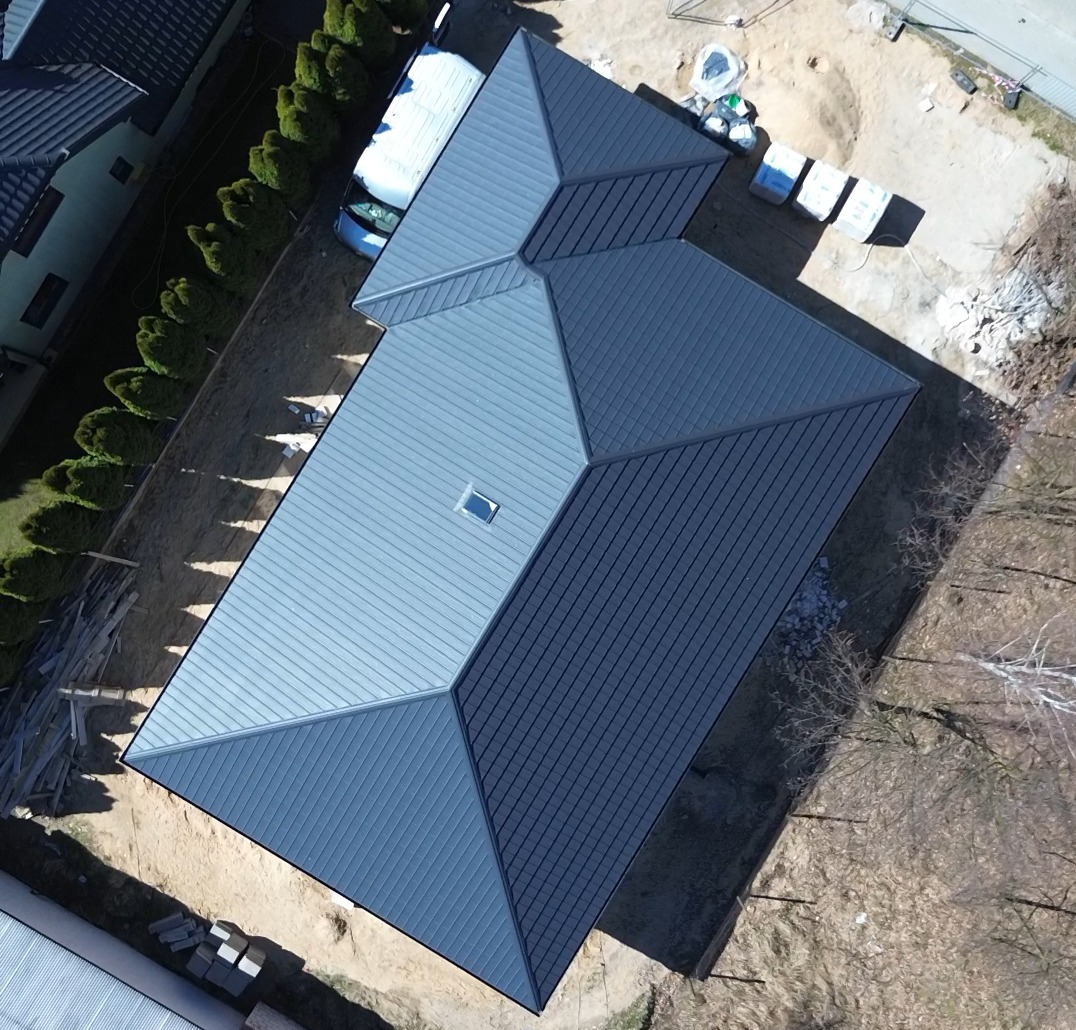 Zdjęcie z drona przedstawia ukończony dach z blachy na domu w stanie deweloperskim. Widoczne elementy konstrukcyjne, okolica i materiały budowlane.