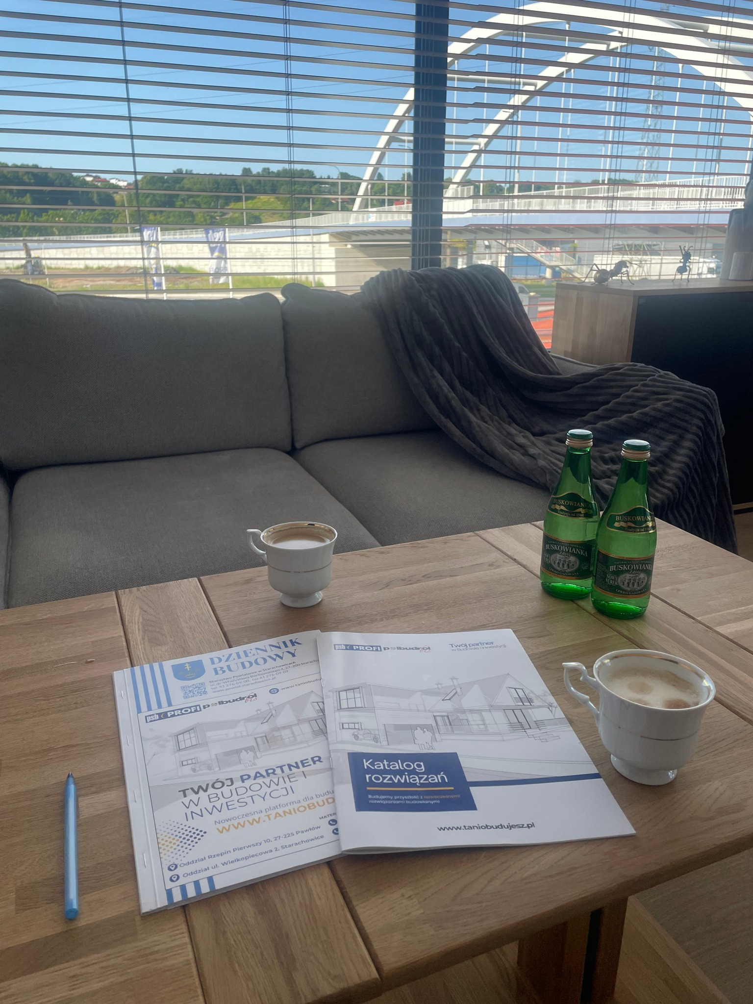 Katalog z wizualizacją domu na stole w jasnym wnętrzu z sofą i widokiem na zewnątrz przez żaluzje. Dwie butelki wody i filiżanki kawy dodają przytulności.