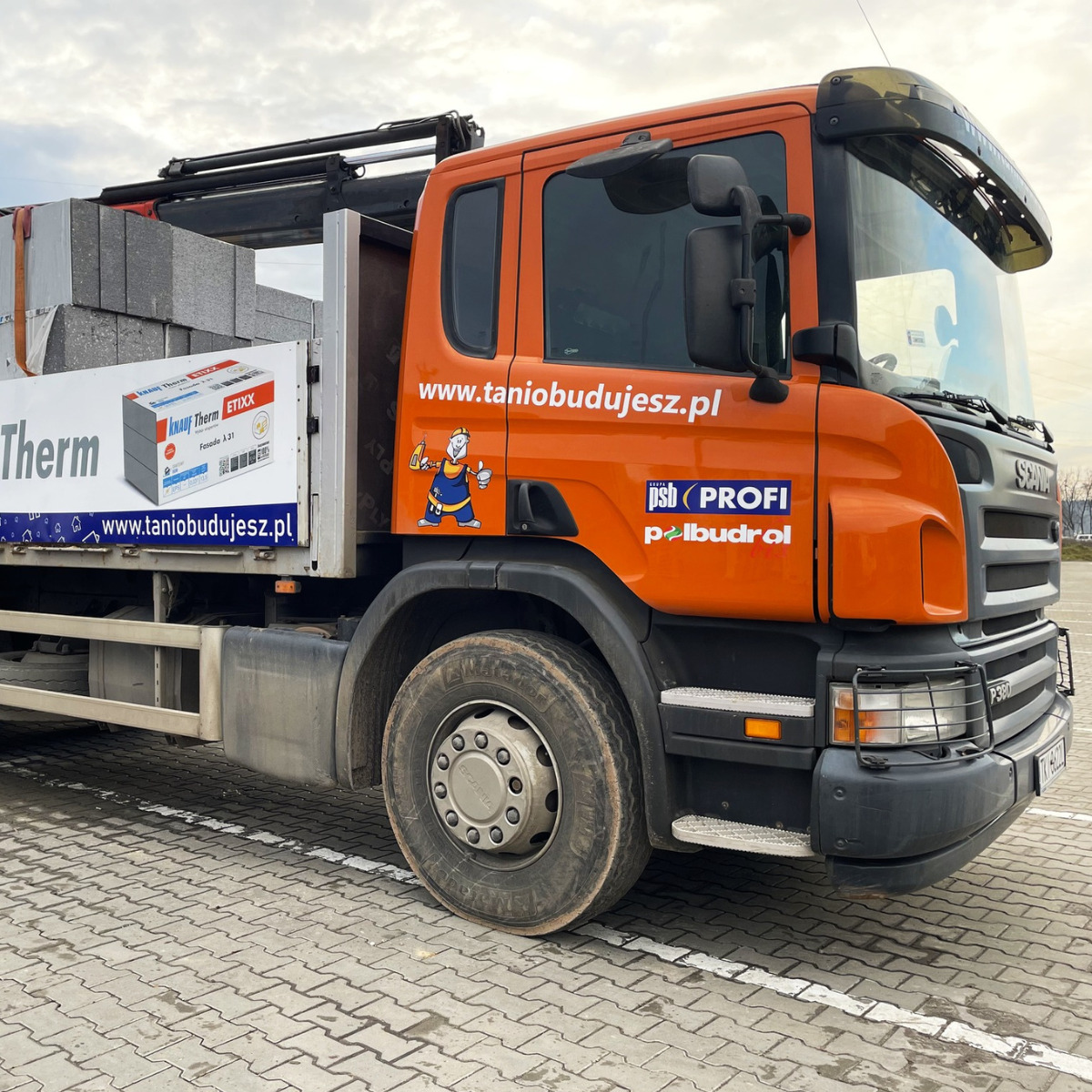 Pomarańczowa ciężarówka Scania z reklamą taniobudujesz.pl, załadowana materiałami budowlanymi Knauf Therm, stoi na kostce brukowej. Widoczny rysunek murarza.