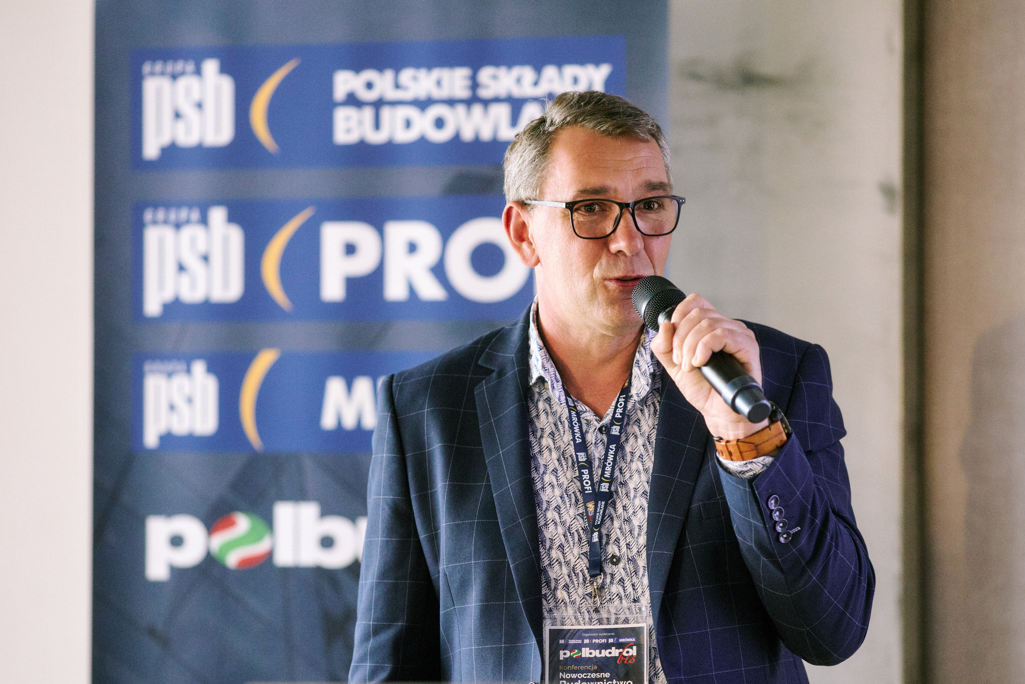Mężczyzna w okularach przemawia do mikrofonu na tle baneru PSB i Polbudrol podczas konferencji Nowoczesne Budownictwo. Widoczny identyfikator.