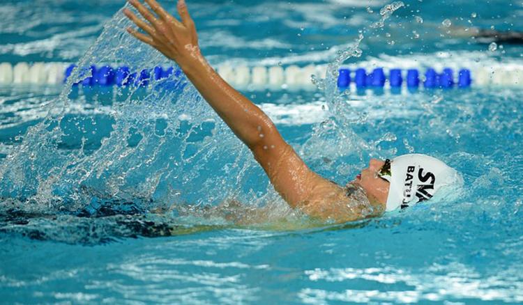 Pływaczka w trakcie treningu, styl grzbietowy, biały czepek z napisem SVA, woda rozpryskuje się wokół ręki, widoczne tory basenowe.