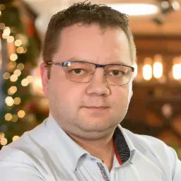 Portret mężczyzny w okularach, w jasnej koszuli, z delikatnym uśmiechem na tle rozmytych światełek. Wyraźne detale koszuli i oprawek okularów.