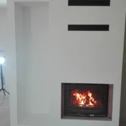 Biała obudowa kominka z wkładem kominkowym i otworami wentylacyjnymi, widoczne kable zwisające z sufitu i lampa budowlana na statywie w tle, surowy beton na podłodze.