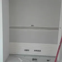 Wnętrze w trakcie wykończenia z kominkiem, ściana przygotowana pod malowanie z widocznymi łączeniami płyt, listwa dekoracyjna na dole ściany, poziomica oparta o ścianę.