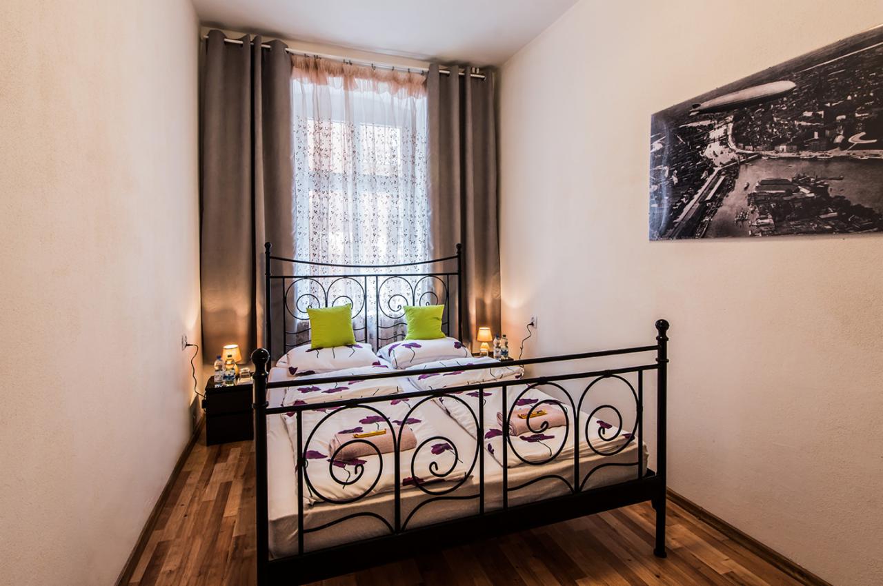 Pokój hotelowy z dwuosobowym łóżkiem z czarną, metalową ramą, zielonymi poduszkami i pościelą w kwiaty, nocnymi szafkami z lampkami i czarno-białym zdjęciem na ścianie.