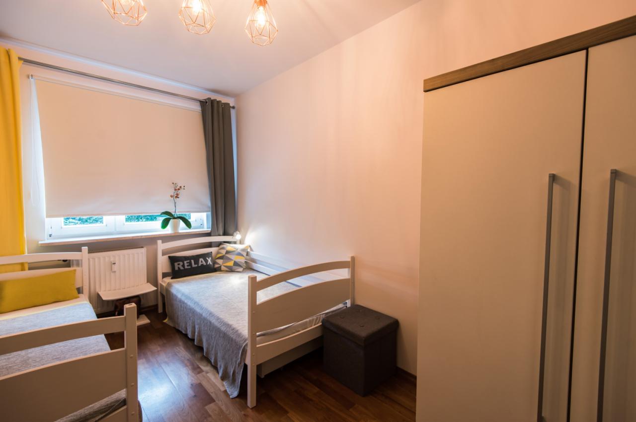 Wnętrze pokoju z dwoma łóżkami, szafą, zasłoniętym oknem i trzema lampami wiszącymi, utrzymane w jasnej kolorystyce.