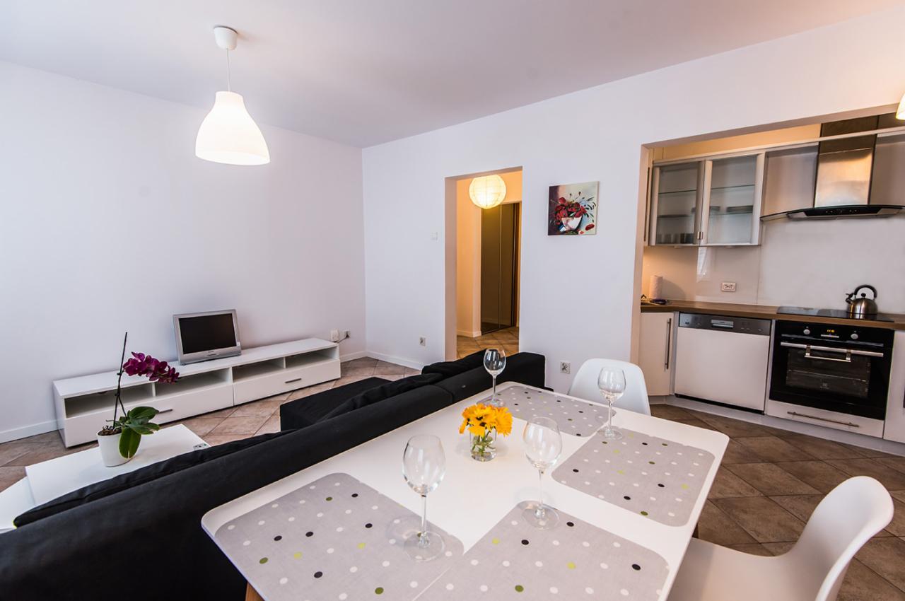 Jasne wnętrze apartamentu z aneksem kuchennym, stołem nakrytym dla czterech osób, czarną sofą i telewizorem na białej szafce RTV, oświetlone lampami wiszącymi.