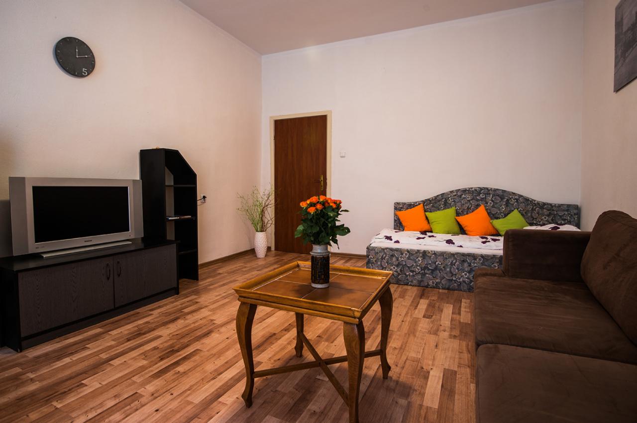 Uporządkowany pokój dzienny z drewnianą podłogą, meblami w ciemnych odcieniach i akcentami kolorystycznymi w postaci pomarańczowych i zielonych poduszek na kanapie, na stoliku bukiet pomarańczowych...