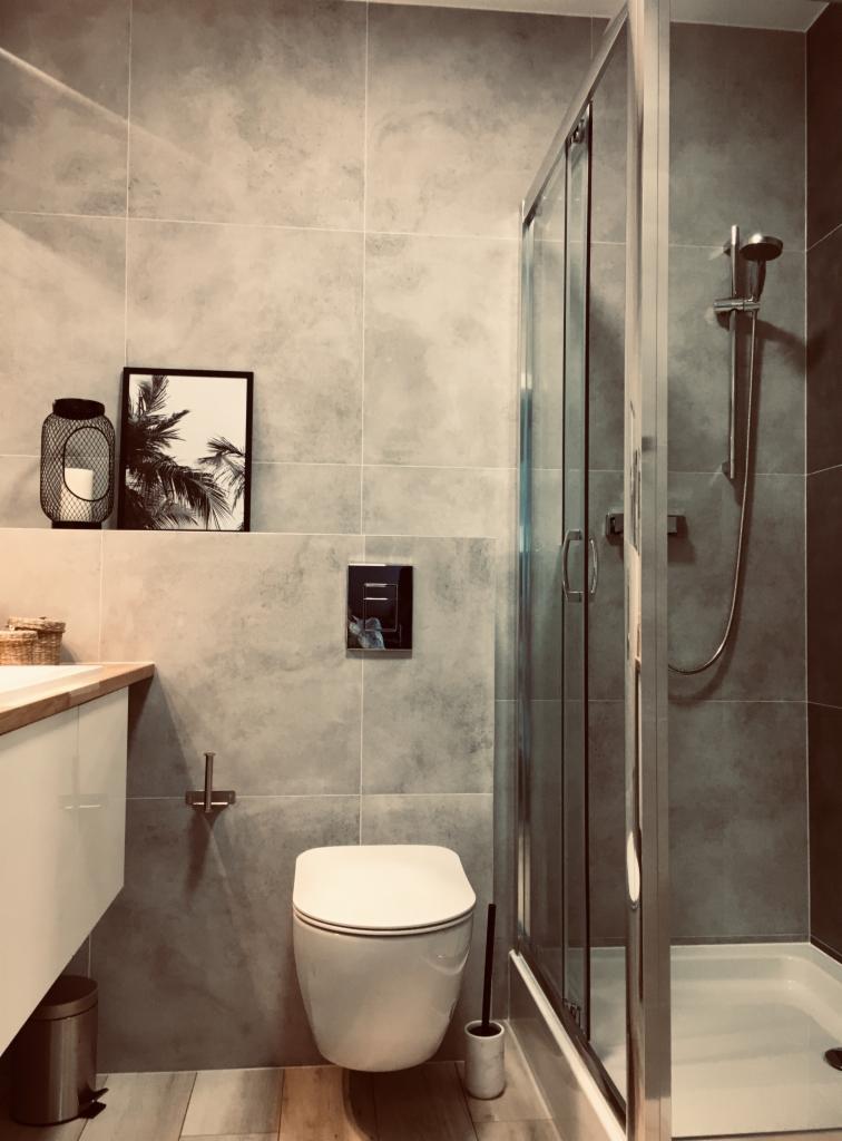 Nowoczesna łazienka w szarościach z kabiną prysznicową, podwieszaną toaletą, umywalką z szafką i akcentami dekoracyjnymi: czarna latarenka i grafika z motywem palm.