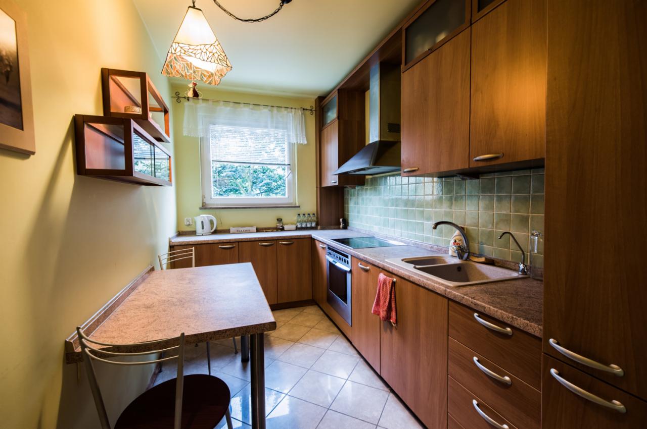 Ustawna kuchnia z drewnianymi szafkami, zielonymi płytkami i stołem przy ścianie, oświetlona lampą z abażurem w stylu vintage.