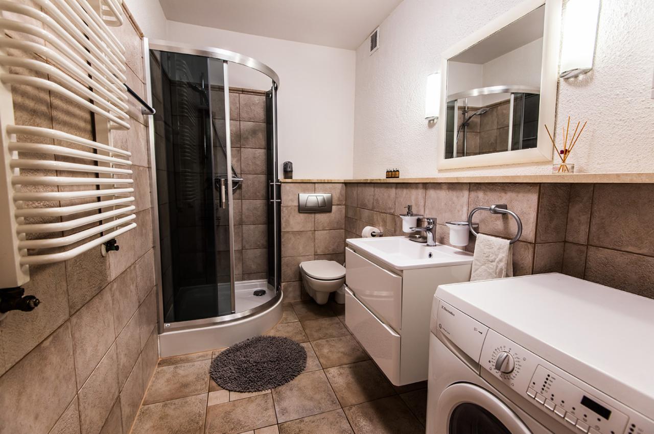 Umywalka z szafką, toaleta, kabina prysznicowa z ciemnymi szybami i pralka w łazience wykończonej brązowymi płytkami.