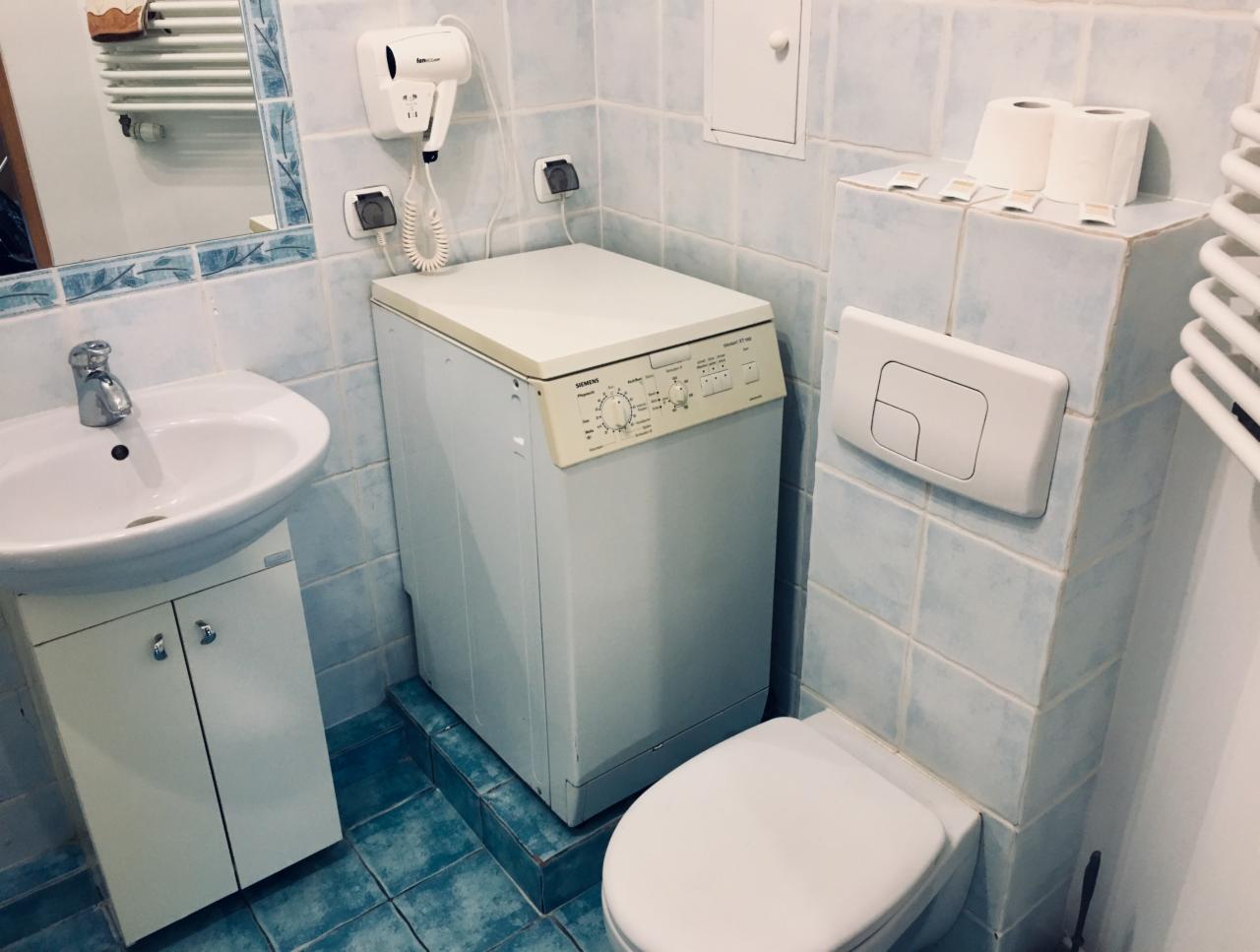 Łazienka z pralką Siemens, umywalką z szafką i toaletą, wykończona niebieskimi płytkami ceramicznymi, z suszarką do włosów na ścianie i grzejnikiem drabinkowym.