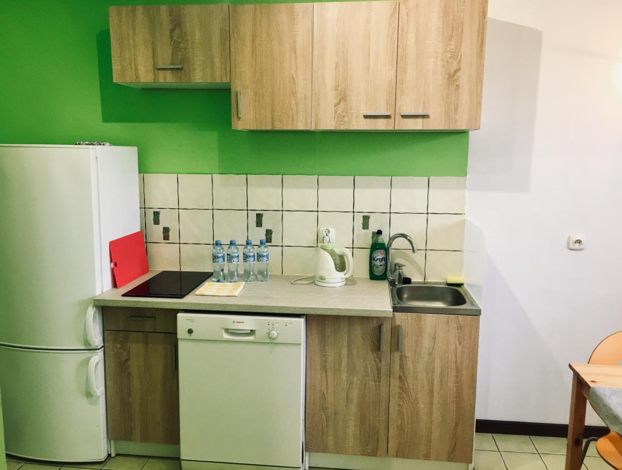 Uporządkowana kuchnia w biurze: biała lodówka, zlew ze środkiem czystości, czajnik elektryczny, butelki wody i szafki z jasnego drewna na tle zielonej ściany.