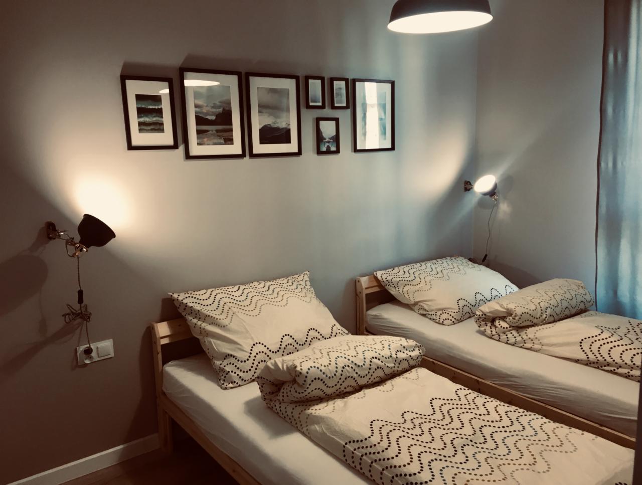 Pokój z dwoma łóżkami pojedynczymi, pościel w zygzakowate wzory, na ścianie galeria obrazków w czarnych ramkach, oświetlenie punktowe z lampkami ściennymi.