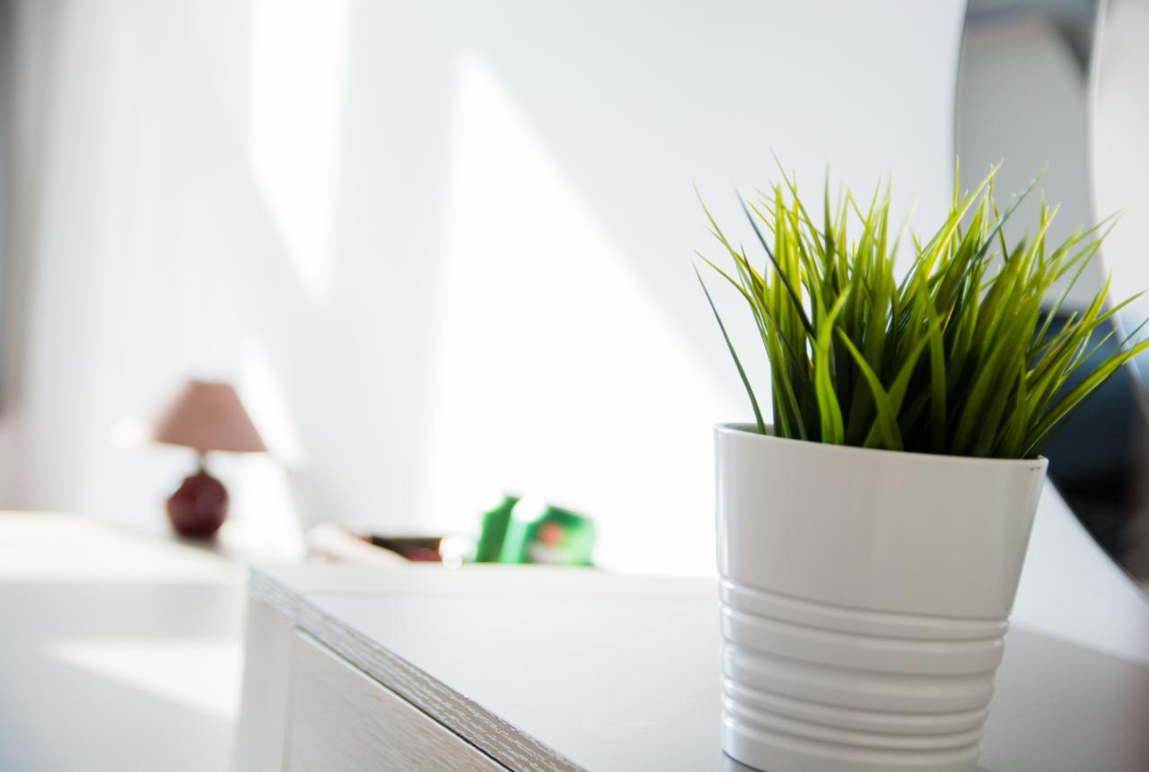 Sztuczna trawa w białej, ceramicznej doniczce z poziomymi żłobieniami, ustawiona na jasnym blacie, w tle rozmyta lampka nocna i biała ściana z refleksami świetlnymi.