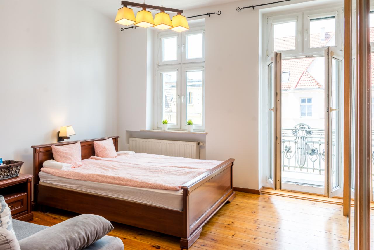 Jasne wnętrze sypialni z drewnianym łóżkiem, różową pościelą i balkonem z widokiem na miasto.