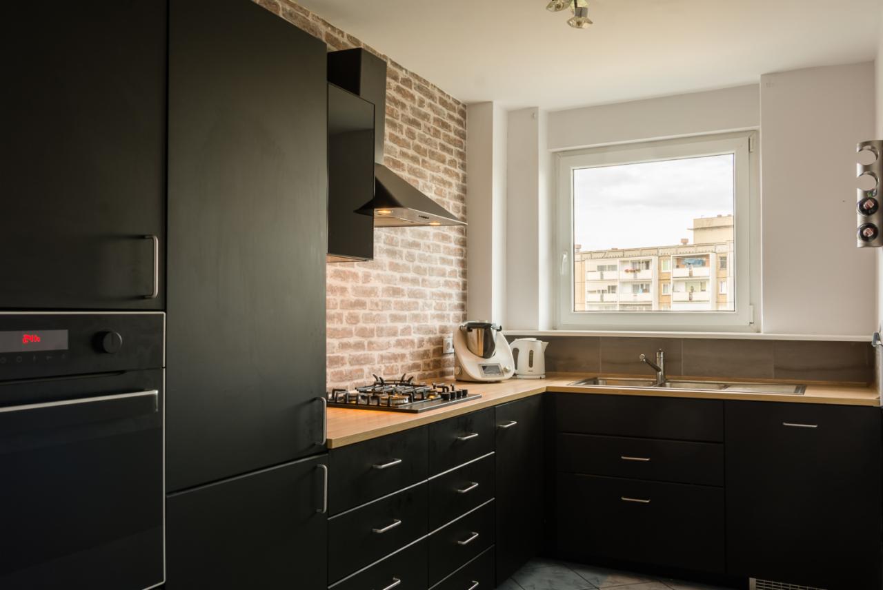 Nowoczesna kuchnia z czarnymi matowymi szafkami, drewnianym blatem i ścianą z cegły, widok na okno i budynek mieszkalny