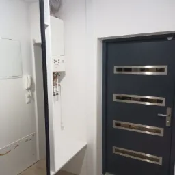 Nowoczesne drzwi wejściowe w kolorze antracytowym z trzema poziomymi, prostokątnymi przeszkleniami ze stali nierdzewnej, widoczne w przedpokoju z białymi ścianami i zabudową z piecem gazowym.