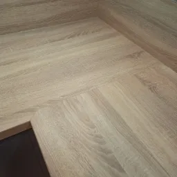 Fragment jasnego blatu z drewnopodobnym wzorem, widoczny narożnik i krawędź, z elementami instalacji elektrycznej w tle.