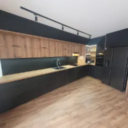 Nowoczesna kuchnia z czarnymi szafkami dolnymi i drewnopodobnymi górnymi, oświetlenie szynowe, czarny zlew i bateria, drewniana podłoga.