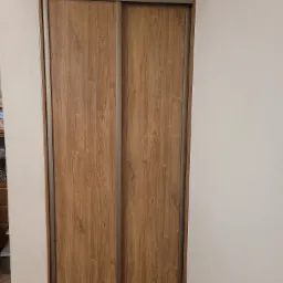 Zamknięta szafa wnękowa z przesuwnymi drzwiami o wzorze jasnego drewna, widoczna z bliska na tle białej ściany.