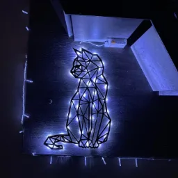 Geometryczny kształt kota, podświetlony niebieskimi diodami LED, umieszczony na ciemnym drewnianym panelu z widocznym zasilaniem bateryjnym i fragmentem zabudowy meblowej.