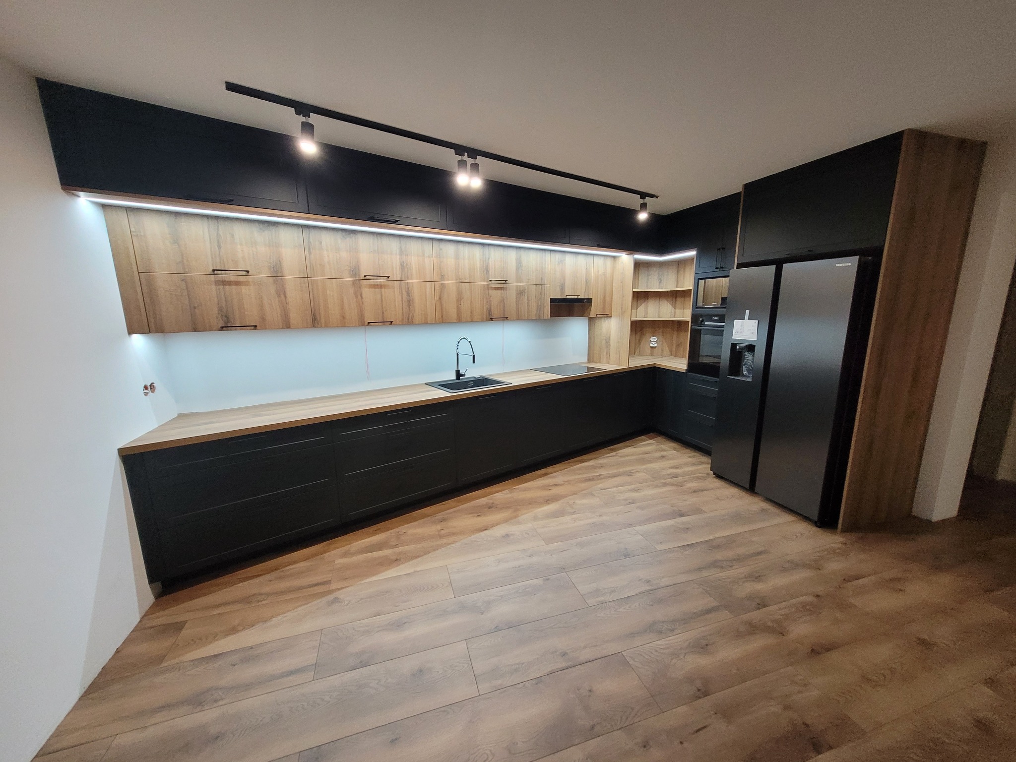 Nowoczesna kuchnia z drewnianymi frontami szafek górnych i czarnymi szafkami dolnymi, oświetleniem punktowym i pod szafkami, z drewnianą podłogą i czarną lodówką.