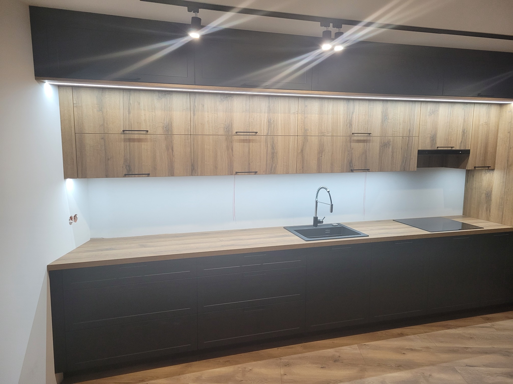 Nowoczesna kuchnia z czarnymi szafkami dolnymi i drewnopodobnymi szafkami górnymi, oświetlona listwą LED pod szafkami wiszącymi i reflektorami sufitowymi. Widoczny zlew z baterią i płyta indukcyjna.