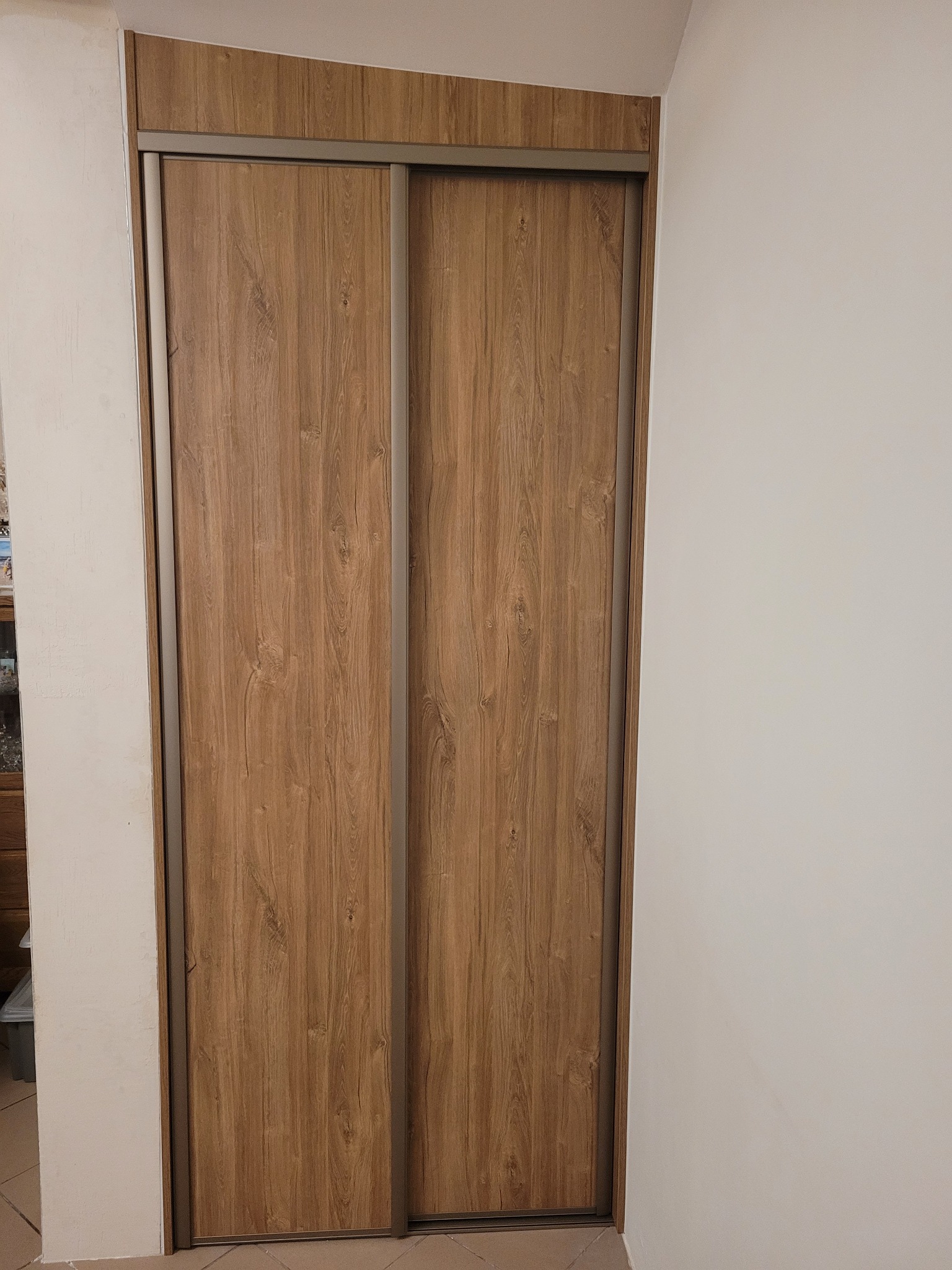 Zamknięta szafa wnękowa z przesuwnymi drzwiami o wzorze jasnego drewna, widoczna z bliska na tle białej ściany.