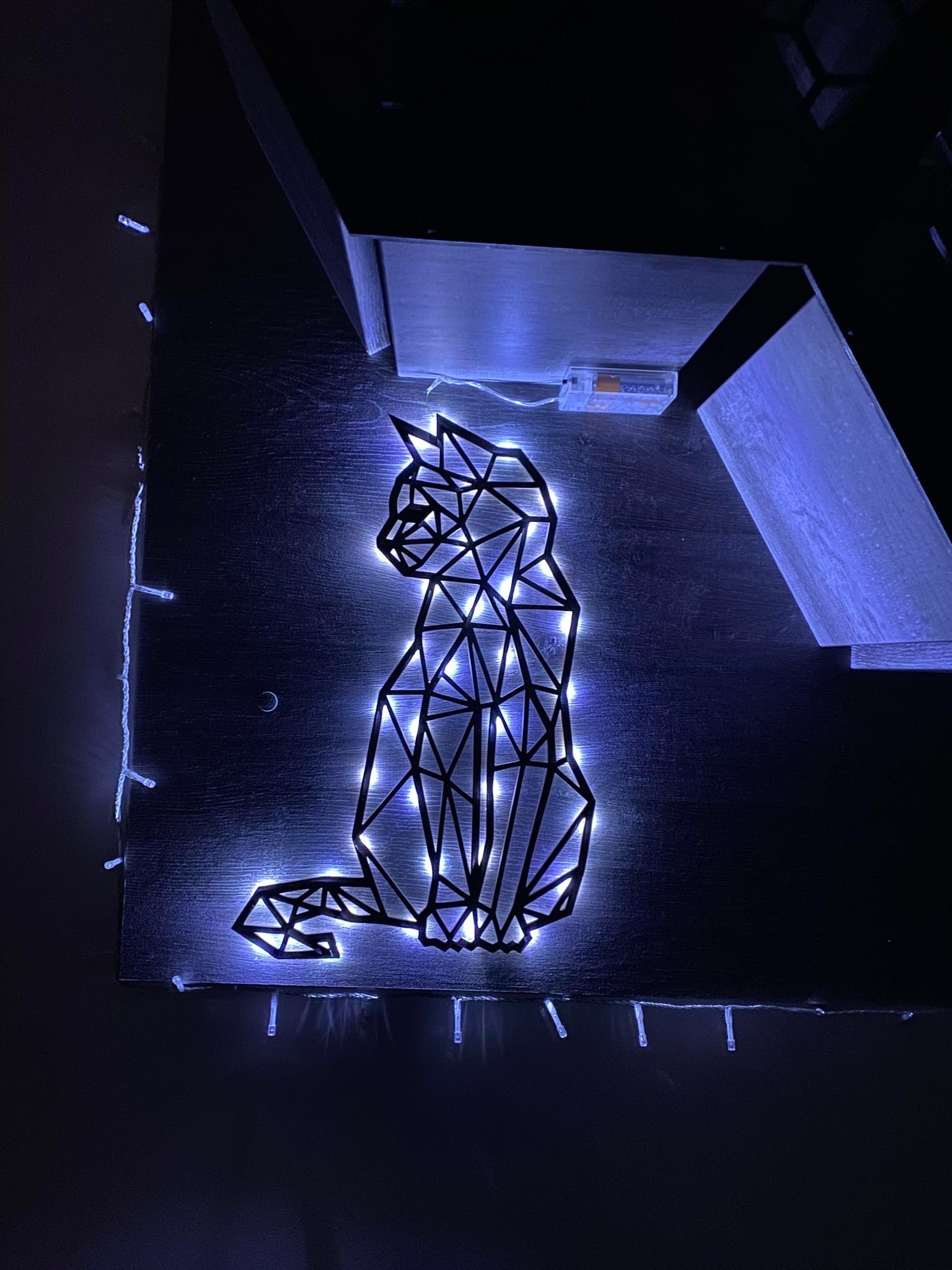 Geometryczny kształt kota, podświetlony niebieskimi diodami LED, umieszczony na ciemnym drewnianym panelu z widocznym zasilaniem bateryjnym i fragmentem zabudowy meblowej.
