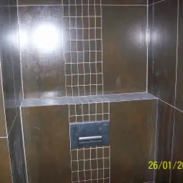 Ciemnobrązowe, błyszczące płytki w łazience ułożone z białymi fugami, z widocznym przyciskiem spłuczki toalety. Fragment ściany wykończony mozaiką z małych kwadratowych płytek.