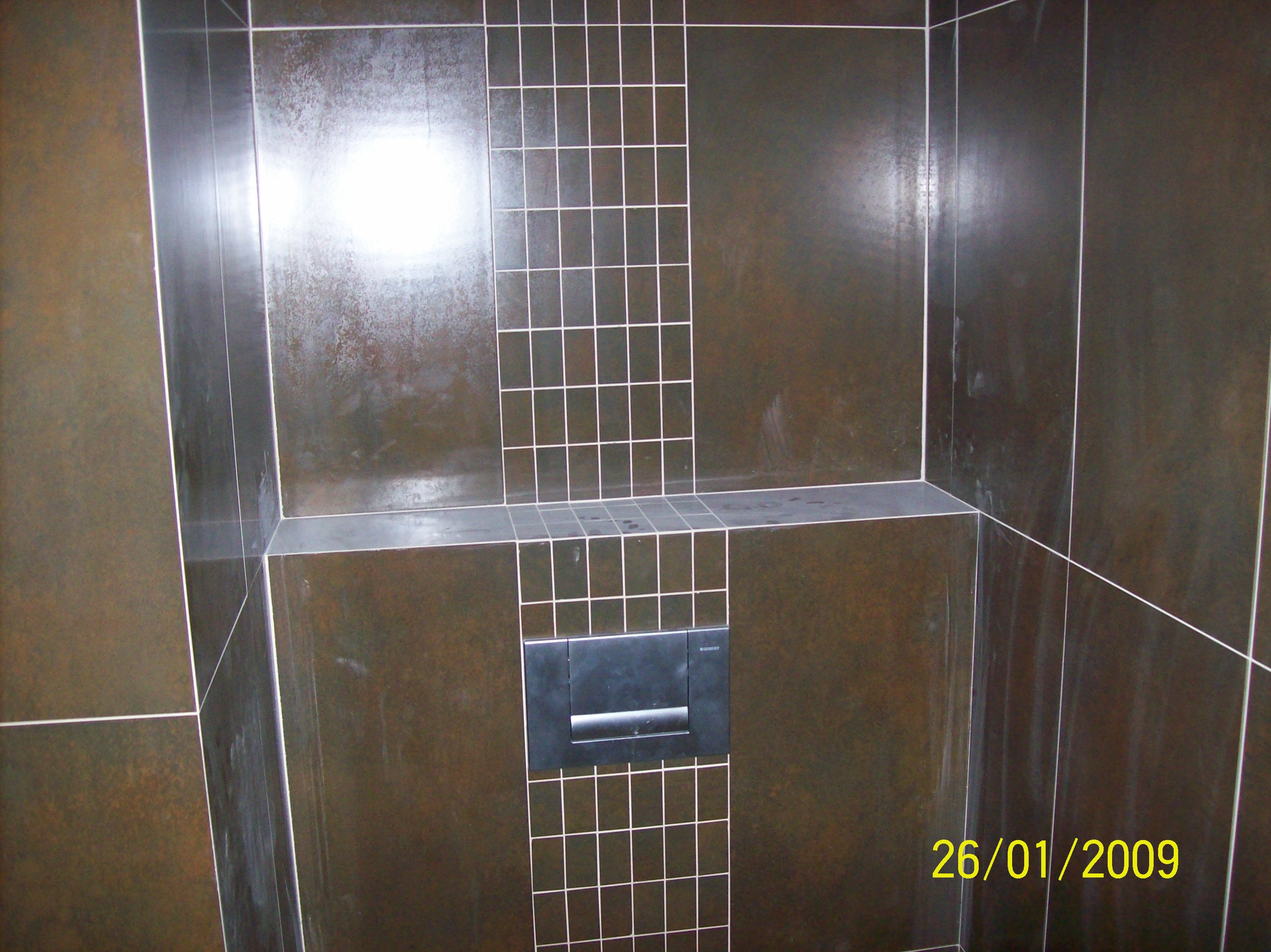 Ciemnobrązowe, błyszczące płytki w łazience ułożone z białymi fugami, z widocznym przyciskiem spłuczki toalety. Fragment ściany wykończony mozaiką z małych kwadratowych płytek.