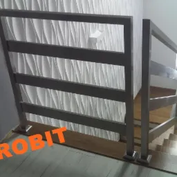 Szara, metalowa balustrada schodowa z czterema poziomymi poprzeczkami, zamontowana na drewnianych schodach. W tle biała ściana z dekoracyjną, falistą strukturą i biały włącznik światła.