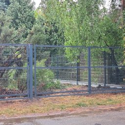 Krzysztof Staroń - Szara, metalowa konstrukcja ogrodzenia z siatką na betonowej podmurówce, widok z zewnątrz posesji, mokry asfalt na pierwszym planie, w tle zieleń drzew i krzewów.