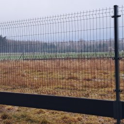 Krzysztof Staroń - Nowoczesne ogrodzenie z siatki z podmurówką betonową, widoczne słupki i przęsła, na tle pola i lasu w pochmurny dzień.